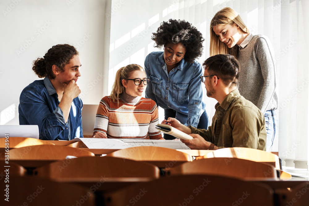 a diverse group of five young adults—three women and two men—gathered around a large table in a sunlit classroom